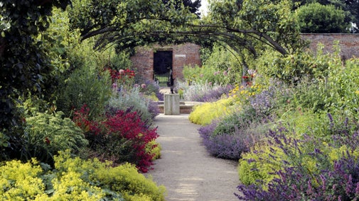 Colourful flower borders spill out over a path leading to a statue and door in a brick wall